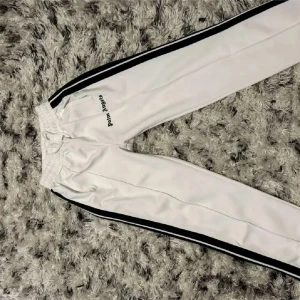 Vita träningsbyxor från Palm Angels - Snygga vita träningsbyxor från Palm Angels med svarta sidoremsor. Byxorna har elastisk midja och en avslappnad passform, perfekt för en sportig look. Märkesloggan är tryckt på framsidan. Priset sänks mycket i snabb affär
