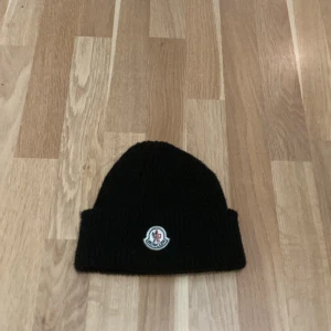 Svart stickad mössa från Moncler - Snygg svart stickad mössa från Moncler med broderad logotyp framtill. Perfekt för att hålla värmen under kyliga dagar. Använd ett få tall gånger men är som ny. Pris diskuteras vid snabb affär.