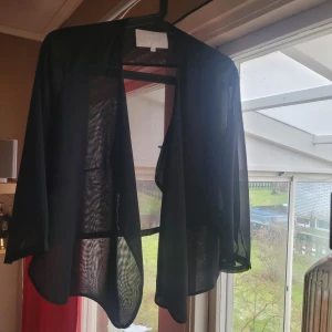 Svart transparent blus från Vila - Säljer en svart transparent blus från Vila i storlek S. Blusen har en luftig och lätt design med trekvartsärmar, perfekt för lager-på-lager-stil. Den är öppen framtill och ger en elegant touch till din outfit. Har släppt lite i en söm som är lätt att fixa, se bild. 