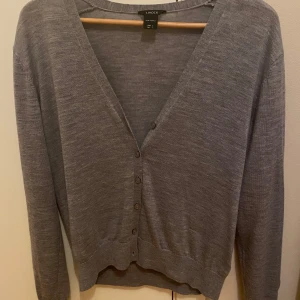 Grå kofta från Lindex i merinoull - Säljer en stilren grå kofta från Lindex i 100% merinoull. Koftan har en v-ringad design med knappar framtill och långa ärmar. Perfekt för att hålla sig varm och snygg under kyligare dagar.