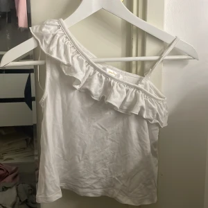 Vit oneshoulder topp från Gina Tricot - Säljer en söt vit oneshoulder topp från Gina Tricot med volangdetaljer längs axeln. Perfekt för varma sommardagar eller en stilren look. Toppen är armlös.🫶🏻