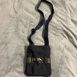 Svart axelväska från Hugo Boss - Snygg svart axelväska från Hugo Boss med justerbar rem och dragkedja. Nypris:1200    Ganska använd men funkar som ny..