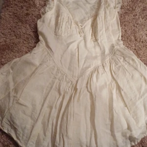 Vit klänning med spetsdetaljer - Charmig vit klänning med vackra spetsdetaljer längs kanterna och en knytning framtill. Klänningen har en luftig passform och är perfekt för varma dagar. Den har en romantisk och vintage-inspirerad stil. Använd en gång 