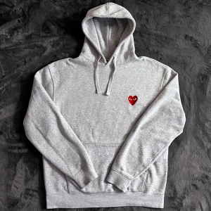 CDG Hoodie - Säljer en stilren grå hoodie från Comme des Garçons med det ikoniska röda hjärtat på bröstet. Tröjan har en klassisk passform med långa ärmar och en praktisk magficka. Perfekt för en avslappnad look. Skick 9/10, Storlek XL men sitter som L. Pris går att diskutera ✅