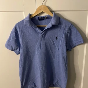 Blå pikétröja från Polo Ralph Lauren - Säljer en klassisk blå pikétröja från Ralph Lauren. Tröjan har korta ärmar och en broderad logotyp på bröstet. Modellen heter slimfit