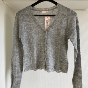 Grå kofta från Nelly M - Säljer en stilren grå kofta från Nelly med knappar framtill. Koftan har en v-ringad design och långa ärmar. Perfekt för lager-på-lager under kyligare dagar.