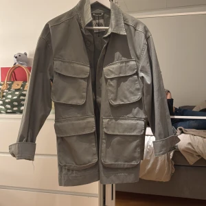 Grön jacka från Zara - Säljer en stilren grå jacka från Zara med flera stora fickor framtill. Jackan har en klassisk krage och knäppning fram. Perfekt för en avslappnad look.