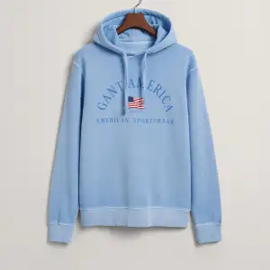 Säljer en snygg ljusblå hoodie från Gant med tryck av amerikansk flagga och texten 'Gant America'. Tröjan har en klassisk känguruficka och justerbar huva med dragsko. Perfekt för en avslappnad stil.