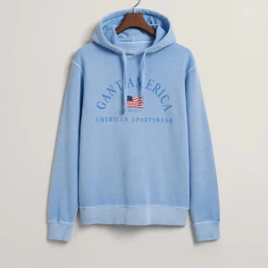 Ljusblå hoodie från Gant - Säljer en snygg ljusblå hoodie från Gant med tryck av amerikansk flagga och texten 'Gant America'. Tröjan har en klassisk känguruficka och justerbar huva med dragsko. Perfekt för en avslappnad stil.