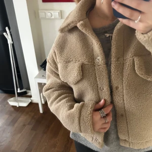 Beige teddyjacka - Mysig beige teddyjacka med knappar framtill och två bröstfickor. Perfekt för kyliga dagar när du vill vara både varm och stilren. Jackan har en klassisk krage och långa ärmar.