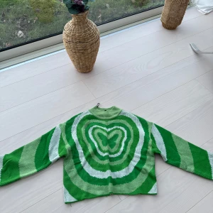 Grön och vit stickad tröja med fjärilsmönster - Säljer en unik grön och vit stickad tröja med ett iögonfallande fjärilsmönster i mitten. Tröjan har långa ärmar och en avslappnad passform. Perfekt för att liva upp garderoben med lite färg och mönster!