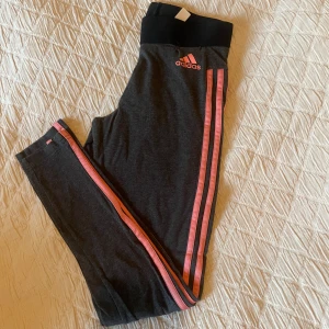 Träningsleggings från Adidas - Träningsleggings från Adidas med rosa detaljer. Mid waist men kan vikas ner till low waist. Har en liten defekt, se bild 2.  