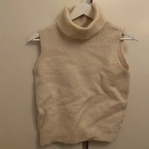 Cremevit stickad polotröja - Säljer en mysig cremevit stickad ärmlös polotröja. Perfekt för lager-på-lager under kyliga dagar. Den har en hög polokrage och är ribbad vid nederkanten.
