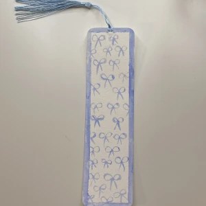 Blå bokmärke med rosett-mönster - Charmigt bokmärke i blått med ett mönster av små rosetter. Det har en matchande blå tofs upptill, perfekt för att hålla koll på var du är i din bok. Stilren och söt design som passar alla bokälskare. Färgen på tofsen går även att bytas ut om så önskas!