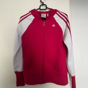 Rosa och vit hoodie från Adidas - Säljer en snygg rosa och vit hoodie från Adidas med dragkedja och huva. Tröjan har långa ärmar med vita detaljer och klassiska ränder längs ärmarna. Perfekt för en sportig look eller avslappnad stil. 🏃‍♀️