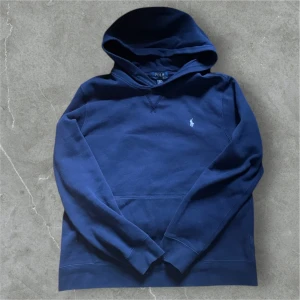 Blå hoodie från Polo Ralph Lauren - Säljer en snygg blå hoodie från Polo Ralph Lauren med det klassiska logotypbroderiet på bröstet. Tröjan har en bekväm passform med långa ärmar och en praktisk huva. Perfekt för en avslappnad stil.