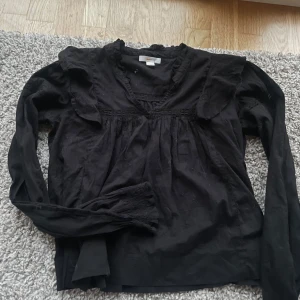 Svart blus med spetsdetaljer - Elegant svart blus i storlek 164/158 med spetsdetaljer och volanger vid axlarna. Blusen har långa ärmar och en v-ringad design som ger en stilren look. Perfekt för att lyfta vilken outfit som helst. Är i fint skick!