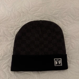  svart mössa med logotyp - Snygg svart mössa med ett diskret rutigt mönster och en logotyp framtill. Perfekt för att hålla värmen under kyliga dagar. Den har en klassisk design som passar till många olika stilar. Inte använd! Helt ny