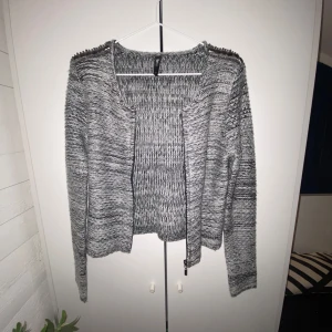 Grå stickad kofta från Flame - Säljer en snygg grå stickad kofta från Flame i storlek S. Koftan har långa ärmar och dekorativa detaljer vid axlarna. Perfekt för kyliga dagar och ger en stilren look.