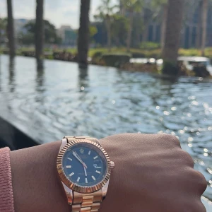 Rose gold day date  - Snygg guldfärgad klocka med svart urtavla och datumvisning. Klockan har en räfflad bezel och en länkarmband i metall. Perfekt för den som vill ha en stilren och elegant accessoar.