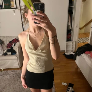 Champagnefärgad topp med kedjedetalj - Snygg champagnefärgad topp med kedjedetalj runt halsen. Toppen har en djup v-ringning och är axelbandslös, vilket ger en elegant look. Perfekt för en utekväll eller festlig tillställning.