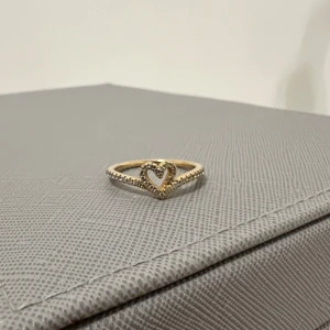 Guldfärgad ring med hjärtdetalj från pandora - Elegant guldfärgad ring med en vacker hjärtdetalj i mitten. Perfekt för att ge en romantisk touch till din stil. Ringen har en smal design som gör den både subtil och stilren. Ringer är från pandora