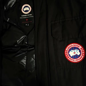 Svart dunjacka från Canada Goose - Säljer en svart dunjacka från Canada Goose med ikonisk logga på bröstet. Jackan har en quiltad insida och dragkedja framtill. Perfekt för kalla vinterdagar med sin varma och skyddande design. Priset kan diskuteras 