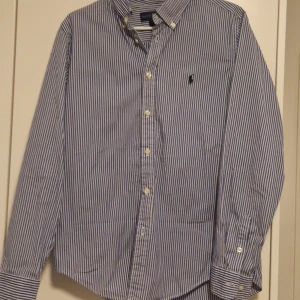 Blå och vit randig skjorta från Ralph Lauren - Snygg blå och vit randig skjorta från Ralph Lauren med broderad logotyp på bröstet. Skjortan har långa ärmar och klassisk krage med knappar. Perfekt för en stilren look. Jag köpte den från en annan men den är tyvärr för liten. Men i absolut toppskick!