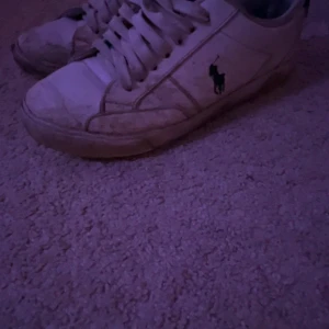 Vita sneakers från Ralph Lauren - Säljer ett par vita sneakers från Ralph Lauren med klassisk snörning och den ikoniska loggan på sidan. Perfekta för en stilren look.