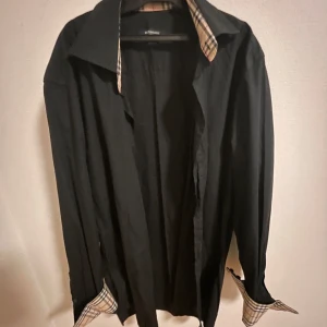 Svart skjorta från Burberry - Snygg svart skjorta från Burberry med klassiska rutiga detaljer på insidan av kragen och ärmsluten. Perfekt för en stilren look. Skjortan har långa ärmar och knappar framtill. Den är i storlek 38 vilket motsvarar S. Hör av er vid frågor och funderingar!