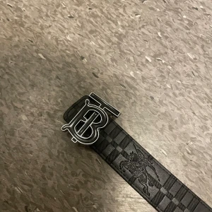 Svart och beige bälte från Burberry - Snyggt bälte  från burberry. Ena sidan är svart med ett diskret rutmönster och den andra sidan har ett klassiskt rutigt mönster i beige och bruna toner. Perfekt för att ge en stilren touch till din outfit.