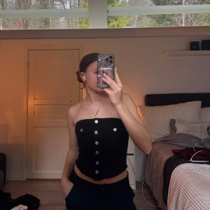 Svart tubtopp med knappar - Snygg svart tubtopp med knappar framtill. Perfekt för en stilren look. Toppen är axelbandslös och har en figurnära passform. Köpte från en tjej på Plick, det står inte vart den är ifrån men skulle gissa SHEIN? Säljer även en i Jeans blå 