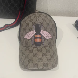 Gucci keps med bi-motiv - Snygg Gucci keps med ett coolt bi-motiv på framsidan. Kepsen har ett klassiskt Gucci-mönster i beige och svart, med en svart mesh-baksida för extra ventilation. Justerbar passform med ett spänne bak. Perfekt för sommardagar när du vill ha en stilren look!