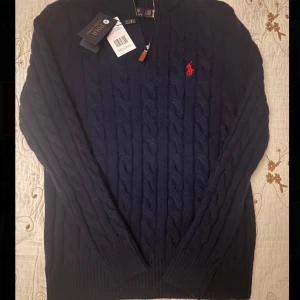 Polo Ralph Lauren - Säljer en snygg mörkblå stickad tröja från Polo Ralph Lauren. Den har ett klassiskt flätmönster och den ikoniska röda loggan på bröstet. Tröjan är långärmad och perfekt för kyliga dagar. Den är i nytt skick med etiketter kvar. Perfekt för både höst och vinter!