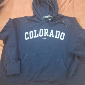 Mörkblå hoodie med text - Säljer en mörkblå hoodie med texten 'COLORADO USA' i vitt på bröstet. Tröjan har en klassisk känguruficka och justerbar huva med dragsko. Perfekt för en avslappnad stil.