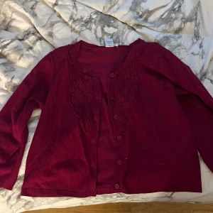 Vinröd kofta med spetsdetaljer - Säljer en mysig vinröd kofta med vackra spetsdetaljer längs halsringningen. Koftan har tre kvarts ärmar och knappar framtill. Perfekt för höst och vinter för en stilren look. Passar både till vardags och festliga tillfällen.