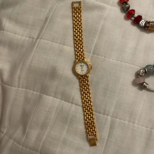 Guldfärgad klocka från Choice - Elegant guldfärgad klocka från Choice med vit urtavla och guldfärgade visare. Armbandet är i en länkdesign som ger en klassisk look. Perfekt för att lägga till en touch av lyx till din stil. Klockan fungerar inte och batteriet måste bytas.