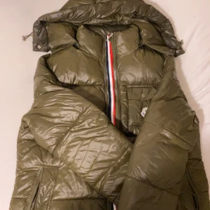 Olive grön pufferjacka från Moncler - Snygg grön pufferjacka från Moncler med ikonisk röd, vit och blå randig dragkedja. Jackan har en praktisk bröstficka med knapp och två sidofickor med dragkedja. Perfekt för kyliga dagar. Jackan är bara 2 veckor gammal köpte den och den var för liten för mig. jackan har en fungerande NFC-tag samt en QR kod på insidan av jackan. Storlek XL men passar M