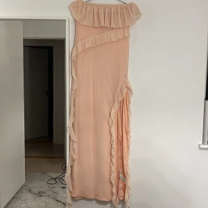 Ljusrosa långklänning med volangdetaljer - Elegant ljusrosa långklänning med volangdetaljer. Klänningen har en offshoulder-design och är dekorerad med volanger längs överdelen och sidan. Perfekt för speciella tillfällen.