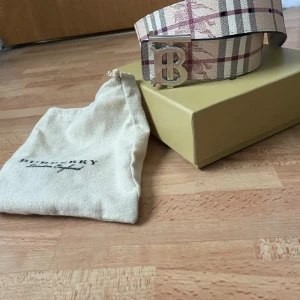 Rutig bälte från Burberry - Snyggt rutig bälte från Burberry med ett stort silverfärgat spänne i form av ett B. Bältet har en klassisk beige och röd färgkombination med Burberrys ikoniska mönster. Perfekt för att ge en stilren touch till din outfit. Bältet är ungefär 112 cm och endast två av hålen har använts men är fortfarande i riktigt bra skick. Hör av dig vid mer frågor! 