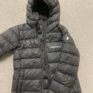 Svart dunjacka från Peak Performance - Säljer en svart dunjacka från Peak Performance med huva och dragkedja. Jackan är perfekt för kalla dagar och har en stilren design med logotyp på bröstet. Storlek xs