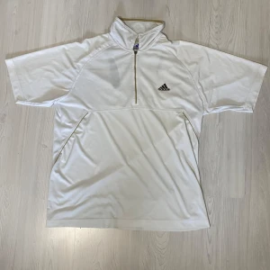 Vit kortärmad tröja från Adidas - Säljer en vit kortärmad tröja från Adidas med dragkedja och klassiska tre ränder på ryggen. Perfekt för sportiga aktiviteter eller en avslappnad stil. Tröjan har en hög krage och är tillverkad i ett lätt och bekvämt material. Några defekter(se bilder). Priset kan diskuteras!