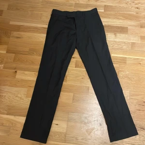 Svarta kostymbyxor från Batistini - Snygga svarta kostymbyxor från Batistini i en mix av polyester och ull. Byxorna har en klassisk straight passform och är perfekta för en stilren look. De har knappar bak och en diskret dragkedja framtill.