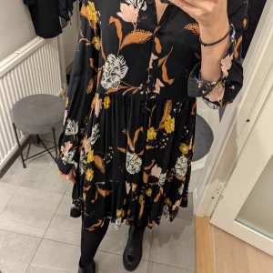 Blommig klänning från Lindex - Säljer en superfin blommig klänning från Lindex i storlek XS. Den är svart med färgglada blommor i rosa, gult och vitt. Klänningen har trekvartsärmar och är gjord i 100% viskos, vilket gör den både bekväm och luftig. 
