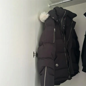 Svart dunjacka från Moose Knuckles - Säljer en svart dunjacka från Moose Knuckles i mycket bra skick. Jackan har en pufferstil med en snygg broderad logga på insidan. Den är perfekt för kalla vinterdagar och har en mysig huva med pälsdetaljer. Dragkedja och knappar framtill för extra värme. Passar både till vardags och festliga tillfällen. Jackan är äkta med 