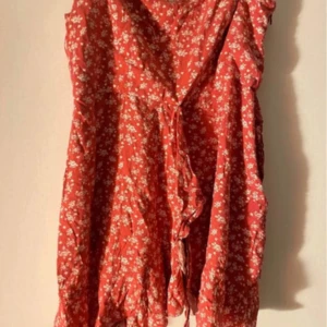 Röd blommig klänning från NA-KD - Säljer en superfin röd klänning med vita blommor från NA-KD. Den har en omlott-design med knytning i midjan och volangdetaljer. Perfekt för sommarens fester eller en mysig dag i solen. Klänningen är i mycket bra skick och gjord av ett lätt och luftigt material.