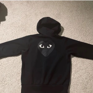 Svart hoodie från Comme des Garçons - Säljer en svart hoodie från Comme des Garçons med det ikoniska hjärtat med ögon på bröstet och ryggen. Perfekt för en stilren och cool look. Hoodien har en normal passform och är i mycket bra skick. Perfekt för höst och vinter!