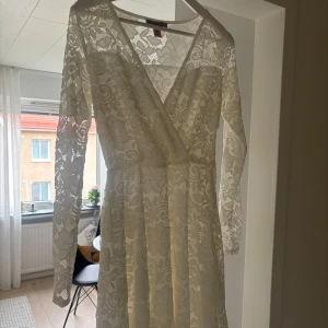 Vit spetsklänning - Elegant vit spetsklänning med omlott-design och v-ringad hals. Klänningen har långa ärmar och ett vackert blommigt mönster. Perfekt för speciella tillfällen. Perfekt till Student och konfirmation