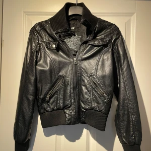 Svart skinnjacka från H&M - Snygg svart skinnjacka från H&M med dragkedja och ribbade muddar. Jackan har två fickor med dragkedjor och en hög krage. Perfekt för en cool look!