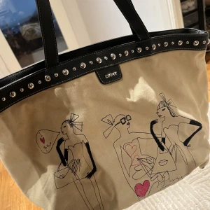Beige shoppingväska från DKNY - Snygg beige shoppingväska från DKNY med svart läderdetaljer och nitar. Väskan har ett unikt tryck av tecknade figurer och hjärtan. Perfekt för en trendig look.